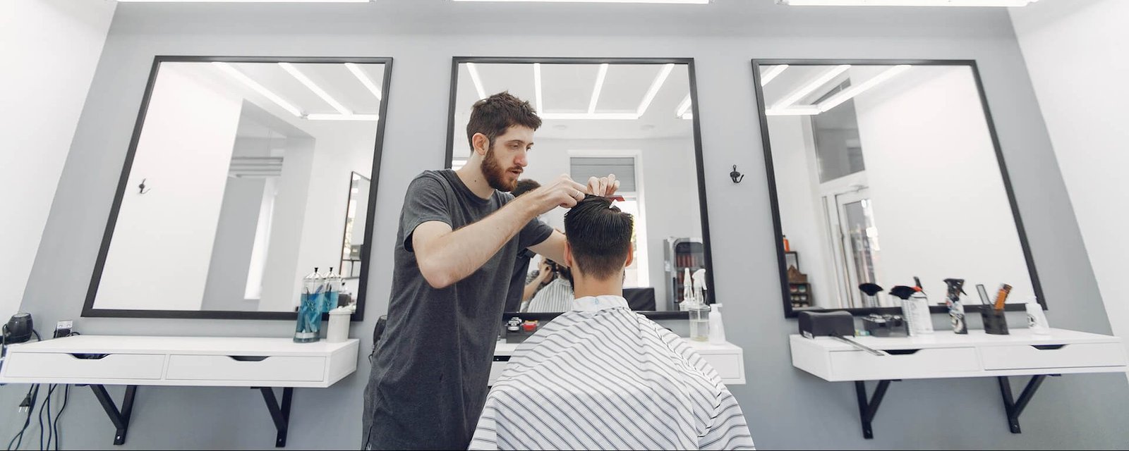 Barbero de barba y camiseta oscura cortando el pelo a un cliente desde la parte trasera en una barbería moderna con decoración blanca y negra, dos espejos grandes y productos de peluquería.