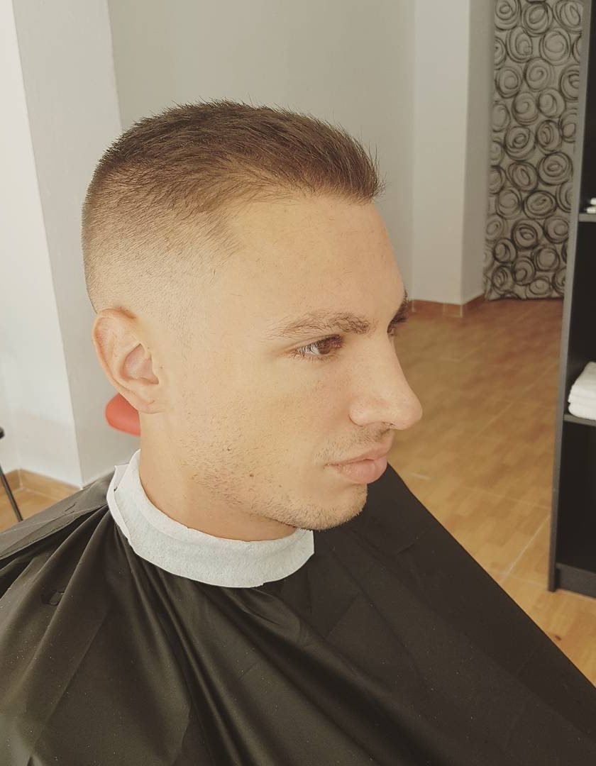 13658466_828758727224140_129187615_n Hombre joven con corte de cabello tipo "fade" muy corto en los lados y ligeramente más largo arriba. Está sentado en la barbería, con fondo de estantería negra y pared blanca.