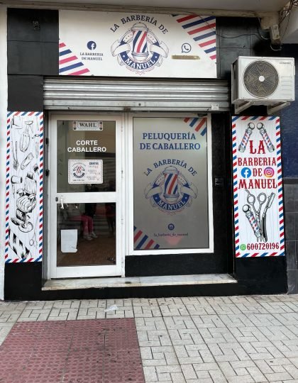 3A35A11DA3CC254FE13C1746001909 Fachada de una barbería llamada "La Barbería de Manuel", con rótulos en los que se leen servicios como "Corte Caballero" y "Peluquería de Caballero". El diseño incluye franjas diagonales en rojo, blanco y azul, típicas de barbería, y hay un aparato de aire acondicionado en la parte superior derecha.