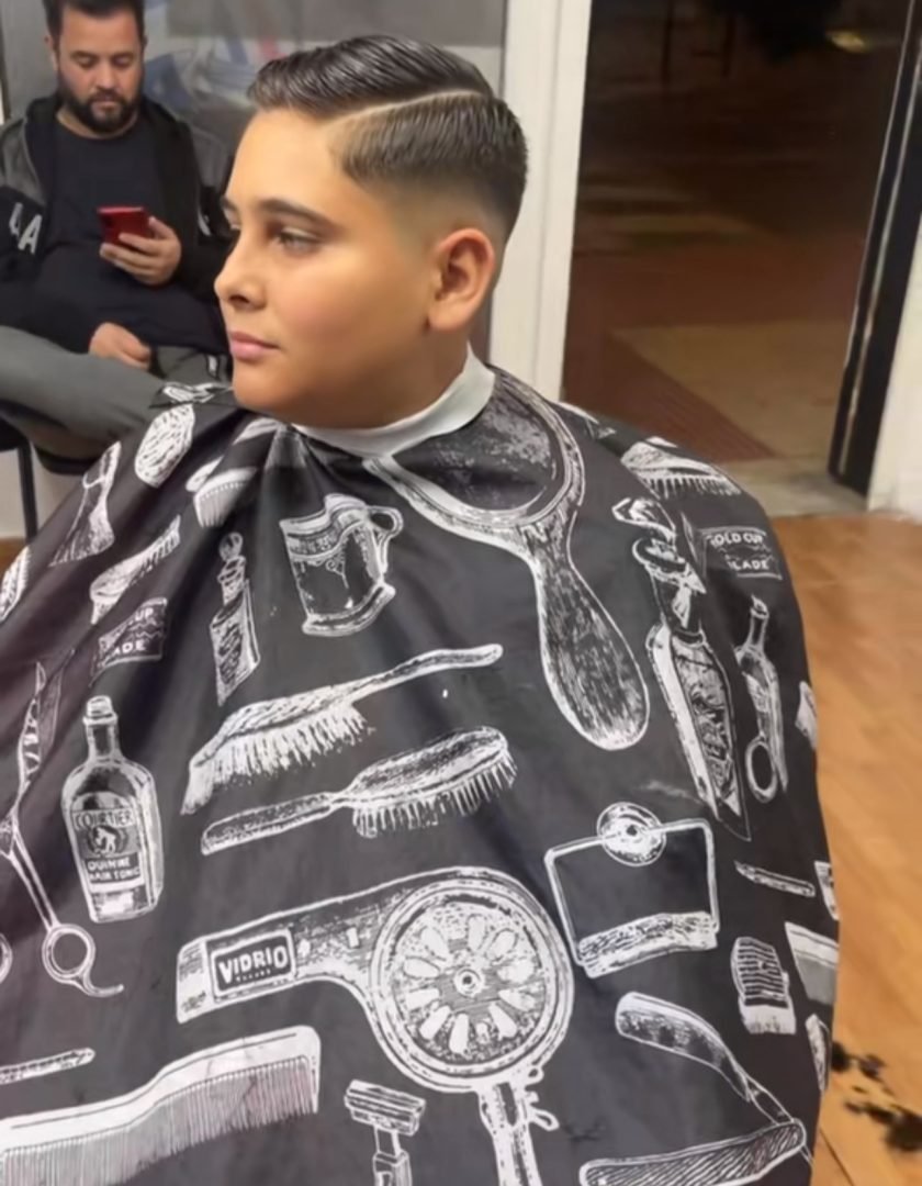 470160983_18348423706120748_2834611916932441024_n Niño sentado en la silla de la barbería, con corte de pelo peinado hacia un lado y línea marcada. Lleva una capa con ilustraciones de herramientas de peluquería.