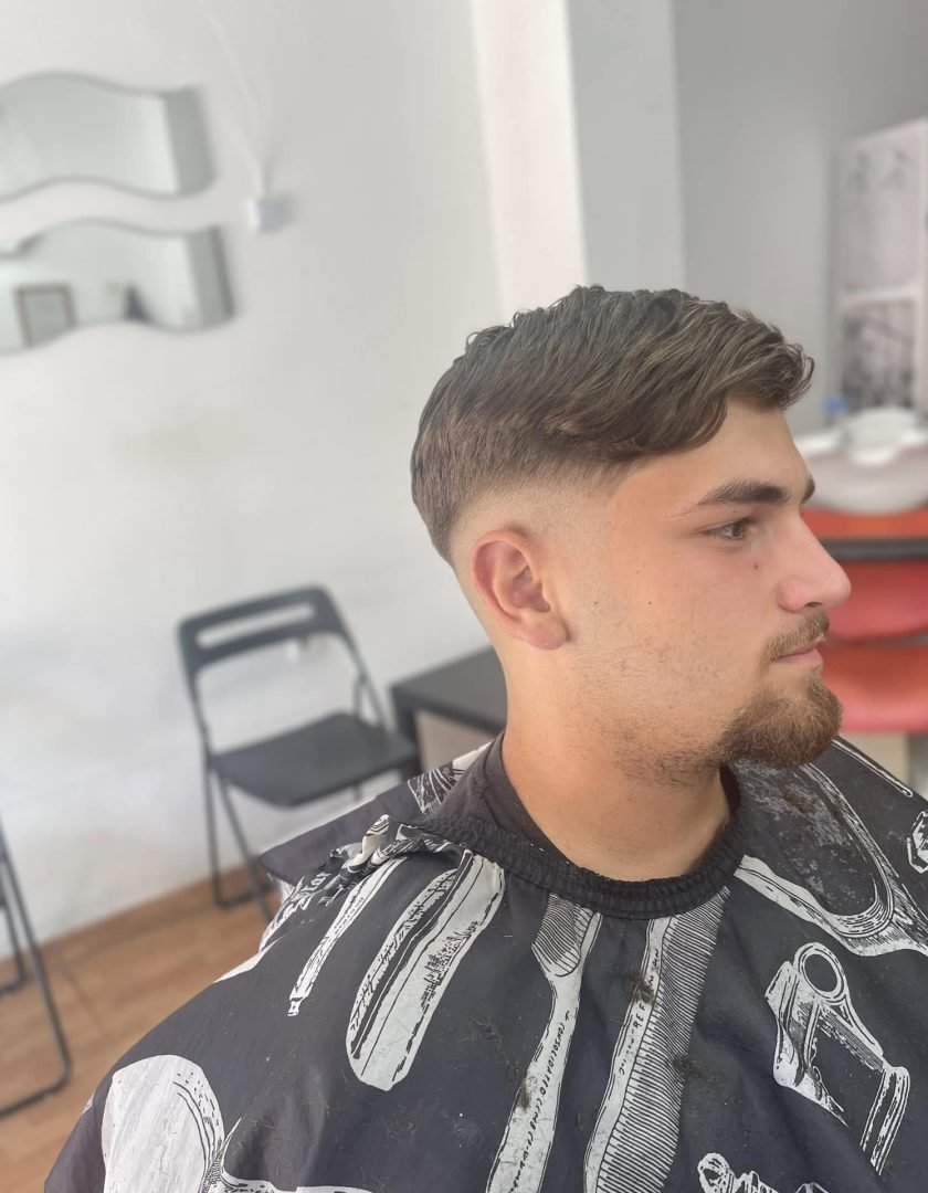 476369397_2054228285015298_1057474000601557860_n Joven con barba corta, mostrando un corte de cabello con degradado alto a los lados y más volumen arriba. Está sentado en la barbería con sillas negras y rojas al fondo.