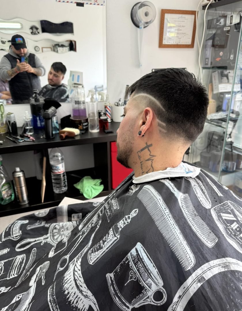 490488583_2109633826141410_3386590510681216716_n Joven con corte de cabello degradado y diseño afeitado en los laterales. Tiene tatuajes en el cuello. Al fondo se ve un espejo y un expositor con productos.